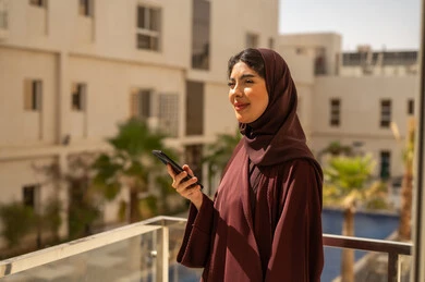 Browsing social media on a mobile phone, having a good time outdoors, an Arab Gulf woman from Saudi Arabia wearing a hijab and an abaya holding a mobile phone in her hands.