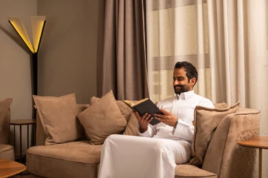 Developing and increasing intellectual resources, a love for reading and studying, an interest in browsing useful books, an Arab Gulf Saudi man wearing a white thobe sitting on the sofa in the living room holding a book in his hand.