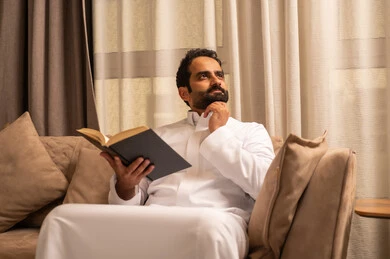 Gestures of confusion and thought, a Saudi Arabian Gulf man wearing a white thobe sits on the sofa in the living room holding a book, a love for reading and literature, development and increase of intellectual wealth.