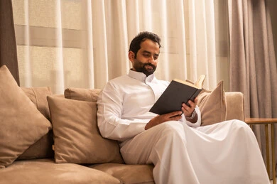 الاهتمام بتصفح الكتب المفيدة، رجل عربي خليجي سعودي يرتدي الثوب الابيض يجلس على الاريكة فى غرفه المعيشة يمسك بيده كتاب ، حب المطالعة والقراءة, تنمية وزيادة الذخيرة الفكرية