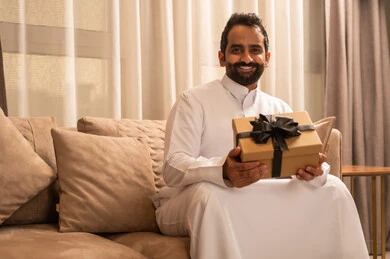 زيادة رابط المحبة بالإهداء والتقدير، تقديم الهدية بالمناسبة العائلية السعيدة,عيد الميلاد والمباركات والأفراح, رجل عربي خليجي سعودي يرتدي الثوب الابيض يجلس على الاريكة فى غرفه المعيشة يمسك بيده علبة هدية ملفوفة بشريط أنيق