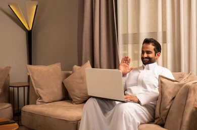 تعابير السلام و التحية ، تطور التكنولوجيا وإستخدام الأجهزة التقنية الحديثة، إنجاز العمل اونلاين من خلال الحاسب المحمول، رجل عربي خليجي سعودي يرتدي الثوب الابيض يجلس على الاريكة فى غرفه المعيشة يقوم بإجراء مكالمة فيديو