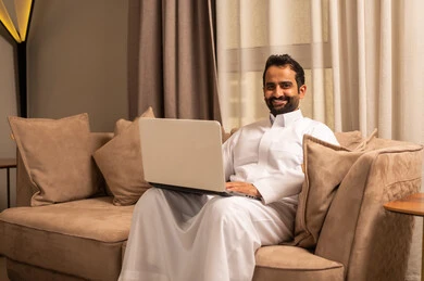 The development of technology and the use of modern technical devices, completing work online through a laptop, a Gulf Arab Saudi man wearing a white thobe sitting on the sofa in the living room making a video call.