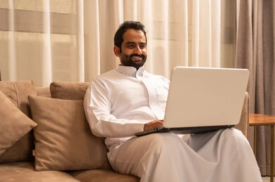 Completing work online through a laptop, an Arab Gulf Saudi man wearing a white thobe sits on the sofa in the living room making a video call, the development of technology and the use of modern technical devices.