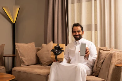 عيد الميلاد والمباركات والأفراح, عرض شاشة سوداء عبر الجوال, زيادة رابط المحبة بالإهداء والتقدير,رجل عربي خليجي سعودي يرتدي الثوب الابيض يجلس على الاريكة فى غرفه المعيشة يمسك بيده علبة هدية ملفوفة بشريط أنيق و هاتف محمول