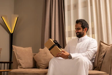 تقديم الهدية بالمناسبة العائلية السعيدة,عيد الميلاد والمباركات والأفراح, زيادة رابط المحبة بالإهداء والتقدير,رجل عربي خليجي سعودي يرتدي الثوب الابيض يجلس على الاريكة فى غرفه المعيشة يمسك بيده علبة هدية ملفوفة بشريط أنيق