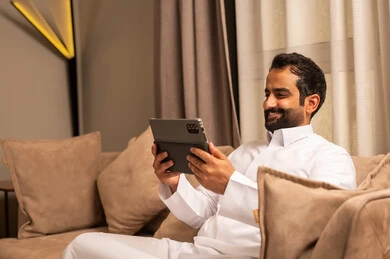 استخدام تطبيق وبرامج الجهاز اللوحي، الدردشة مع الأهل والأصدقاء، استعمال التكنولوجيا و التقنية الحديثة، رجل عربي خليجي سعودي يرتدي الثوب الابيض يجلس على الاريكة فى غرفه المعيشة يحمل بيده جهاز التابلت، تصفح مواقع التواصل الاجتماعي