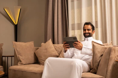 الدردشة مع الأهل والأصدقاء، استعمال التكنولوجيا و التقنية الحديثة، رجل عربي خليجي سعودي يرتدي الثوب الابيض يجلس على الاريكة فى غرفه المعيشة يحمل بيده جهاز التابلت، تصفح مواقع التواصل الاجتماعي، استخدام تطبيقات وبرامج الجهاز اللوحي
