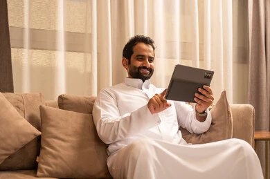 The use of modern technology and techniques, chatting with family and friends, an Arab Gulf Saudi man wearing a white thobe sitting on the sofa in the living room holding a tablet in his hand, browsing social media, using applications and programs on the tablet.