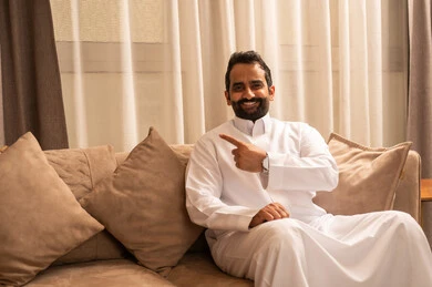 Pointing to an element, clarifying and indicating a specific location, identifying the location of a certain thing, an Arab Gulf Saudi man wearing a white thobe sitting on the sofa in the living room pointing with his index finger forward.