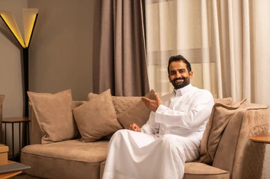 التوضيح والدلالة لمكان محدد، تحديد موقع شيء معين, رجل عربي خليجي سعودي يرتدي الثوب الابيض يجلس على الاريكة فى غرفه المعيشة يشير بيدية للأمام، الاشارة إلى عنصر ما