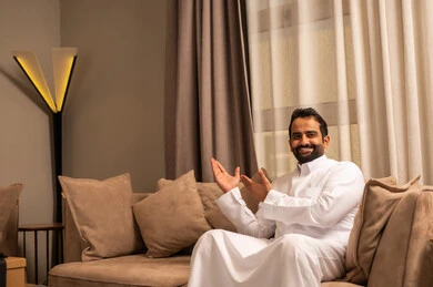 الإشارة إلى عنصر ما, التوضيح والدلالة لمكان محدد,رجل عربي خليجي سعودي يرتدي الثوب الابيض على الاريكة فى غرفه المعيشة يشير بيدية على الجانب، تحديد موقع شيء معين، ايماءات السعادة و السرور