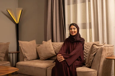 مفهوم الأناقة والأنوثة,امرأة عربية خليجية سعودية ترتدي العباءة والحجاب تجلس على الاريكة فى بايدي متشابكة غرفه المعيشة, التقاط صور شخصية للوثائق الرسمية، النظر إلى الكاميرا بتعابير السعادة والسرور، الاهتمام بالمظهر الخارجي