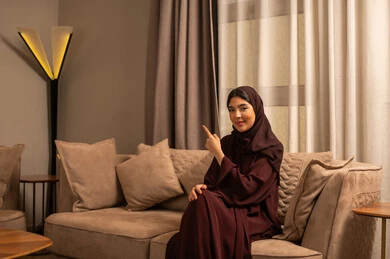 تحديد موقع شيء معين, التوضيح والدلالة لمكان محدد, امرأة عربية خليجية سعودية ترتدي العباءة والحجاب تجلس على الاريكة فى غرفه المعيشة تشير بإصبع السبابة للأمام، الاشارة إلى عنصر ما