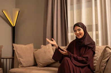 التوضيح والدلالة لمكان محدد, الإشارة إلى عنصر ما, امرأة عربية خليجية سعودية ترتدي العباءة والحجاب تجلس على الاريكة فى غرفه المعيشة تشير بيديها على الجانب، تحديد موقع شيء معين، ايماءات السعادة و السرور