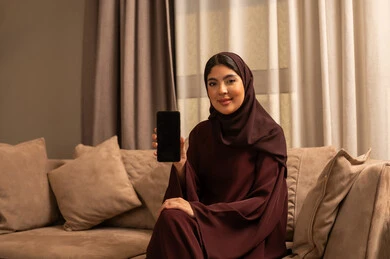 Looking at the camera with gestures of happiness and joy, an Arab Gulf Saudi woman wearing a hijab and an abaya sits on the sofa in the living room displaying a blank black screen through her mobile phone, using modern and advanced devices and technologies.
