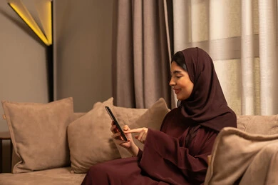 تصفح موقع التواصل الاجتماعي، امراة عربية خليجية سعودية محجبة ترتدي العباءة تجلس على الاريكة فى غرفه المعيشة تحمل بيدها الهاتف المحمول تنظر  بإيماءات السرور، قضاء وقت ممتع عبر الجوال