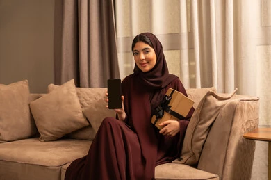 A black screen displayed on the mobile phone, giving gifts on happy occasions, celebrating a birthday, an Arab Gulf Saudi woman wearing a hijab and an abaya sitting on the sofa in the living room holding a gift box and a mobile phone, looking with gestures of happiness and joy.