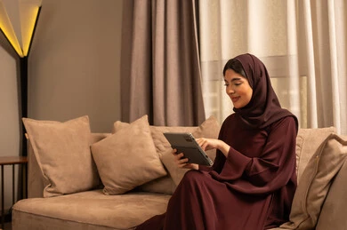 مشاهدة االفيلم و المسلسل المفضل، إنجاز مهام العمل عن بعد، مراقبة مواقع التواصل الاجتماعي، تطور الأجهزة والتقنيات الحديثة، البحث عن شيء ما عبر الانترنت، امرأة عربية خليجية سعودية تجلس على أريكة في غرفة المعيشة و تمسك بيدها جهاز التابليت