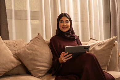 إنجاز مهام العمل عن بعد، مراقبة مواقع التواصل الاجتماعي، تطور الأجهزة والتقنيات الحديثة, مشاهدة الأفلام والمسلسلات المفضلة، البحث عن شيء ما عبر الانترنت، امرأة عربية خليجية سعودية تجلس على أريكة في غرفة المعيشة و تمسك بيدها جهاز التابليت