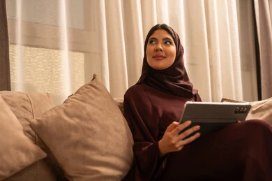 مراقبة مواقع التواصل الاجتماعي، تطور الأجهزة والتقنيات الحديثة, إنجاز مهام العمل عن بعد, مشاهدة الأفلام والمسلسلات المفضلة، البحث عن شيء ما عبر الانترنت، امرأة عربية خليجية سعودية تجلس على أريكة في غرفة المعيشة و تمسك بيدها جهاز التابليت