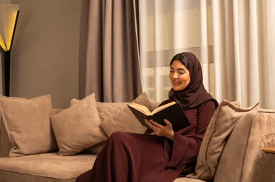 ايماءات التركيز والاستمتاع، تمضية أوقات الفراغ بالتعلم والتثقيف الذاتي، امراة عربية سعودية خليجية ترتدي العباءة و الحجب تجلس على الاريكة في غرفه المعيشة تحمل بيدها كتاب, قضاء أوقات هادئة في المنزل, قراءة الروايات والقصص