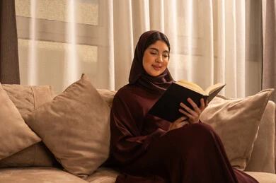Spending a quiet time at home, using free time for learning and self-education, a Saudi Arabian Gulf woman wearing an abaya and hijab sits on the sofa in the living room holding a book, reading novels and stories, gestures of focus and enjoyment.
