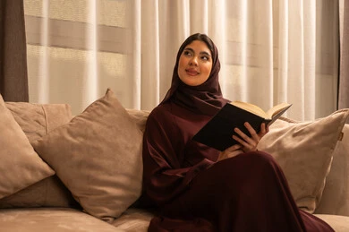 النظر الى شيء ما، قضاء وقت هاديء في المنزل، تمضية أوقات الفراغ بالتعلم والتثقيف الذاتي، امراة عربية سعودية خليجية ترتدي العباءة و الحجب تجلس على الاريكة في غرفه المعيشة تحمل بيدها كتاب, قراءة الروايات والقصص, ايماءات التركيز والاستمتاع