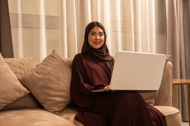 تصفح الانترنت واستخدام تقنية حديثة، إجراء مكالمة فيديو، الدردشة و التواصل الاجتماعي , العمل عن بعد, امراة عربية خلجية سعودية ترتدي العباءة و الحجاب تجلس في غرفة المعيشة, استخدام الحاسوب المحمول