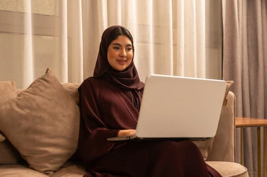 الدردشة و التواصل الاجتماعي, إجراء مكالمة فيديو , العمل عن بعد, امراة عربية خلجية سعودية ترتدي العباءة و الحجاب تجلس في غرفة المعيشة, استخدام الحاسوب المحمول, تصفح الانترنت واستخدام تقنيات حديثة