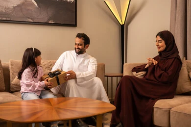 جو عائلي سعيد، تبادل الحديث والدردشة مع الأبناء, التهنئة بقدوم العيد ,اب  عربي خليجي سعودي يرتدي الثوب الابيض يجلس في غرفه المعيشة برفقة زوجته يقوم بتقديم الهدايا لابنته في الأعياد والمناسبات, ايماءات الفرح بالمفاجآت
