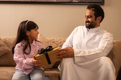 Talking and chatting with children, congratulating on the arrival of Eid, an Arab Gulf Saudi father wearing a white thobe sitting in the living room presenting gifts to his daughter during holidays and occasions, gestures of joy with surprises, happy family atmosphere.