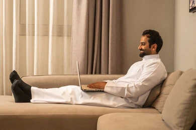Remote work, typing on a keyboard, using a modern and advanced technical device to complete projects, an Arab Gulf Saudi man wearing a white thobe sitting on the sofa in the living room using a laptop with gestures of joy.
