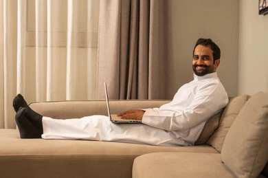 Using a modern and advanced technical device to complete projects, working remotely, typing on the keyboard, an Arab Gulf Saudi man wearing a white thobe sitting on the sofa in the living room using a laptop with gestures of joy.