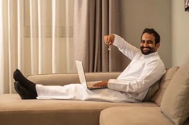 انجاز العمل والمهام عن بعد عبر الحاسوب المحمول، ايماءات وجه تدل على السعادة بإمتلاك منزل جديد، تصفح الانترنت ووسائل التواصل الاجتماعي، رجل عربي خليجي سعودي يرتدي الثوب الابيض يجلس على الاريكة فى غرفه المعيشة يحمل بيديه مفاتيح