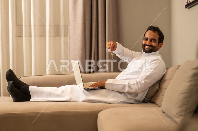 ايماءات وجه تدل على السعادة بإمتلاك منزل جديد، انجاز العمل والمهام عن بعد عبر الحاسوب المحمول، تصفح الانترنت ووسائل التواصل الاجتماعي، رجل عربي خليجي سعودي يرتدي الثوب الابيض يجلس على الاريكة فى غرفه المعيشة يحمل بيديه مفاتيح
