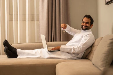 ايماءات وجه تدل على السعادة بإمتلاك منزل جديد، انجاز العمل والمهام عن بعد عبر الحاسوب المحمول، تصفح الانترنت ووسائل التواصل الاجتماعي، رجل عربي خليجي سعودي يرتدي الثوب الابيض يجلس على الاريكة فى غرفه المعيشة يحمل بيديه مفاتيح