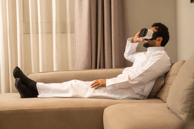 Spending free time in an enjoyable way, watching a virtual reality movie, using advanced technology and tech, immersing and interacting with the imaginary world, an Arab Gulf Saudi man wearing a white thobe sitting on the sofa in the living room wearing VR glasses on his eyes.