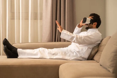 مشاهدة فيلم الواقع الافتراضي، استخدام التقنية والتكنولوجيا المتطورة، الاندماج والتفاعل مع العالم الخيالي، رجل  عربي خليجي سعودي يرتدي الثوب الابيض يجلس على الاريكة فى غرفه المعيشة يضع نظارة في آر على عينيه، قضاء أوقات الفراغ بشكل ممتع