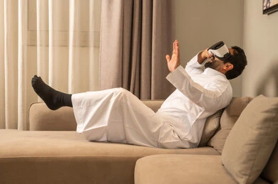 استخدام التقنية والتكنولوجيا المتطورة، الاندماج والتفاعل مع العالم الخيالي، رجل  عربي خليجي سعودي يرتدي الثوب الابيض يجلس على الاريكة فى غرفه المعيشة يضع نظارة في آر على عينيه، مشاهدة أفلام الواقع الافتراضي، قضاء أوقات الفراغ بشكل ممتع