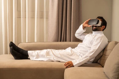 Immersion and interaction with the imaginary world, using advanced technology and tech, an Arab Gulf Saudi man wearing a white thobe sitting on the sofa in the living room wearing VR glasses on his eyes, watching virtual reality movies, spending leisure time in an enjoyable way.
