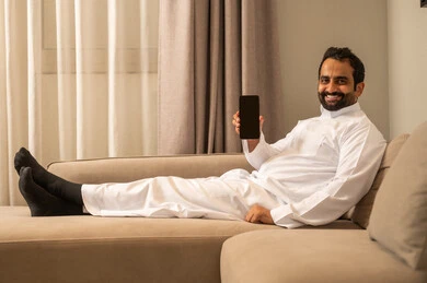 A black screen is displayed on the mobile phone, a Saudi Arabian Gulf man wearing traditional attire is sitting on the sofa in the living room holding a mobile phone in his hand, using a modern and advanced technological device, with expressions of happiness and joy.