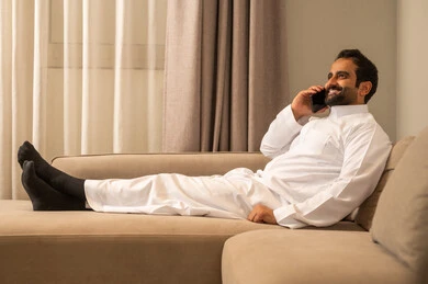Making a phone call via mobile, an Arab Gulf Saudi man wearing traditional attire sits on the sofa in the living room holding a mobile phone in his hand, chatting and communicating with family and friends, using a modern and advanced technological device, expressions of happiness and joy.
