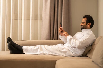 تصفح موقع التواصل الاجتماعي، رجل عربي خليجي سعودي يرتدي الثوب التقليدي يجلس على الاريكة في غرفه المعيشة يمسك بيده الهاتف المحمول، الدردشة والتواصل مع الأهل و الأصدقاء، استخدام جهاز تقني حديث ومتطور، العمل عن بعد وانجاز المهام المطلوبة