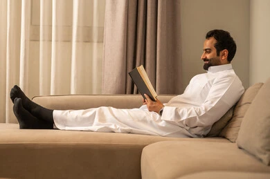 The culture of self-learning, harmony, calmness, and tranquility, enjoying reading a book, a smiling Arab Gulf Saudi man wearing a white thobe holding a book in his hand, gestures of happiness and joy, spending enjoyable time.