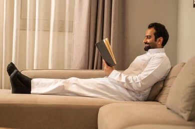 Spending enjoyable time, harmony, tranquility, and serenity, the culture of self-learning, enjoying reading a book, a smiling Arab Gulf Saudi man wearing a white thobe holding a book in his hand, gestures of happiness and joy.