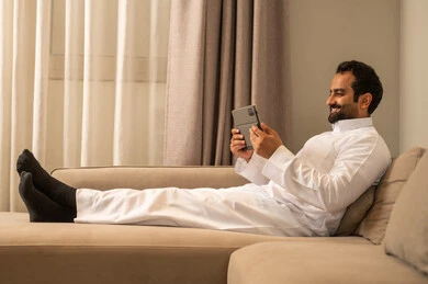 ايماءات السعادة و السرور، استخدام التكنولوجيا و التقنية الحديثة، تصفح موقع التواصل الاجتماعي، متابعة الأفلام والمسلسلات الممتعة، المحادثة مع الأهل والأصدقاء، رجل عربي خليجي سعودي يرتدي الثوب الابيض يجلس على الاريكة فى غرفه المعيشة يحمل بيده جهاز التابلت