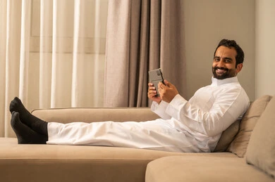 The use of modern technology and techniques, browsing social media, watching enjoyable movies and series, chatting with family and friends, an Arab Gulf Saudi man wearing a white thobe sitting on the sofa in the living room holding a tablet in his hand, gestures of happiness and joy.