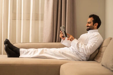 تصفح موقع التواصل الاجتماعي, استخدام التكنولوجيا والتقنيات الحديثة، متابعة الأفلام والمسلسلات الممتعة، المحادثة مع الأهل والأصدقاء، رجل عربي خليجي سعودي يرتدي الثوب الابيض يجلس على الاريكة فى غرفه المعيشة يحمل بيده جهاز التابلت, ايماءات السعادة و السرور