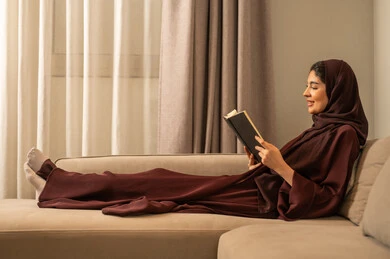 Sitting in the living room, spending time reading and learning, the culture of reading and knowledge, an Arab Gulf Saudi woman wearing a hijab and an abaya sits on the sofa and is reading a book.